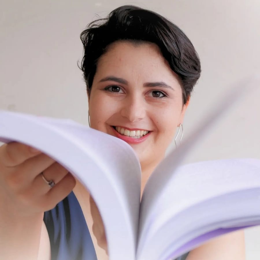 Kássia Magalhães sorrindo enquanto folheia um livro aberto em primeiro plano