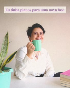 Kássia sorrindo com uma caneca verde nas mãos, sentada à mesa ao lado de livros e de uma planta