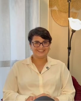 Kássia de óculos e camisa clara, sorrindo em uma sala com luminária e decoração em palha