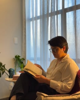 Kássia lendo um livro sentada em uma poltrona, perto de uma janela com cortinas claras