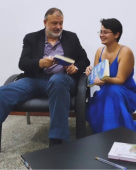 Kássia de vestido azul conversando com um homem de terno, os dois segurando livros