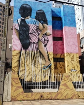 Mural pintado na lateral de um prédio, com uma mulher de costas carregando uma criança